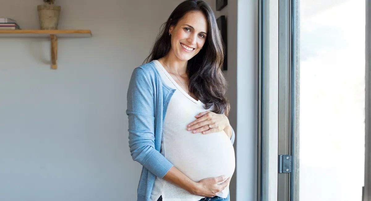 خانمی باردار در حال لمس شکم خود و لبخند زدن کنار پنجره