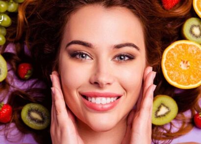 خانمی در حال نشان دادن پوست صورت خود و مقدار زیادی میوه مفید برای پوست