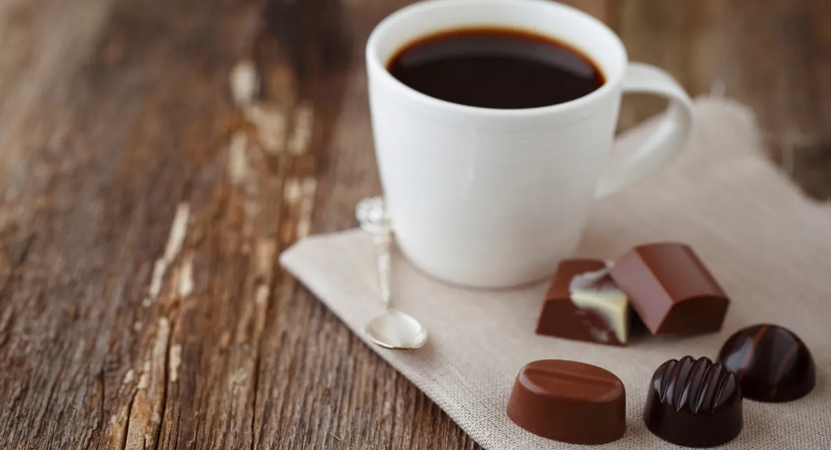 یک فنجان سفید رنگ پر از قهوه به همراه چند تکه شکلات روی یک تکه پارچه سفید