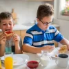 دو پسر بچه در حال خوردن صبحانه پشت میز و آب پرتقال و نان تست روی میز