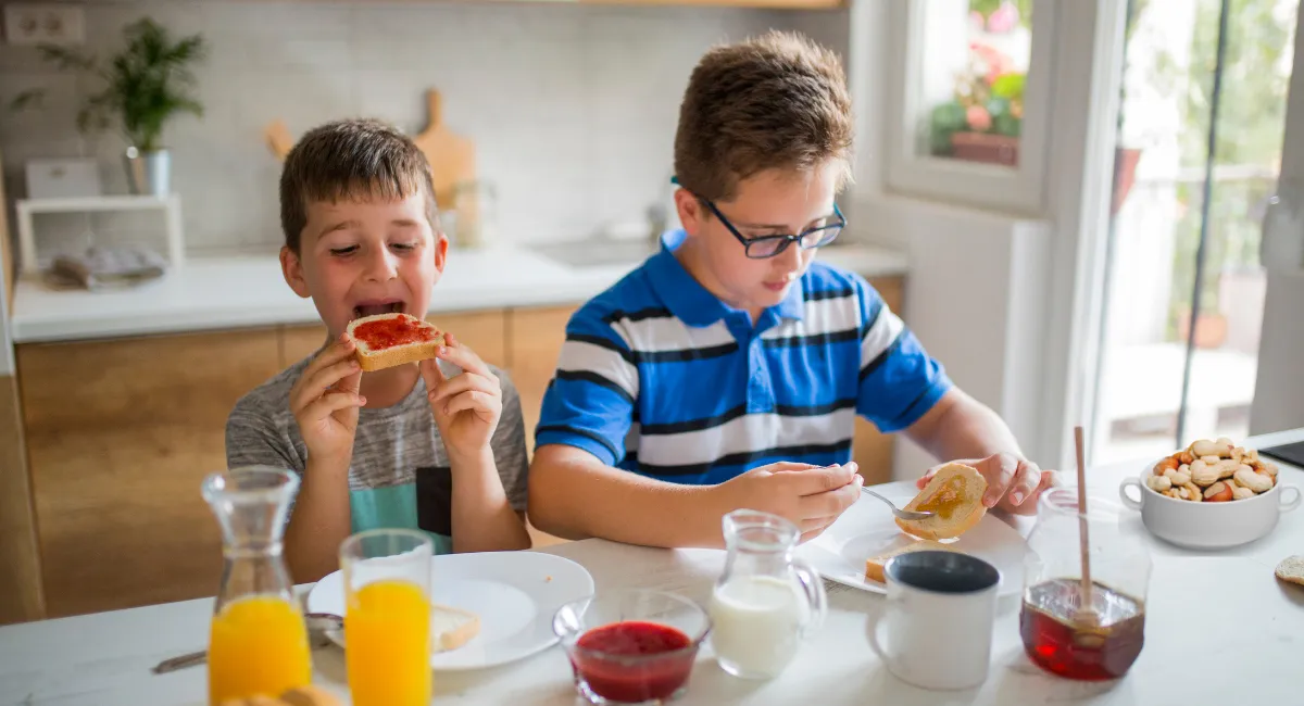 دو پسر بچه در حال خوردن صبحانه پشت میز و آب پرتقال و نان تست روی میز