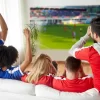 عکسی از پشت سر چند دختر و پسر در حال تماشای فوتبال در تلویزیون و نشسته روی مبل