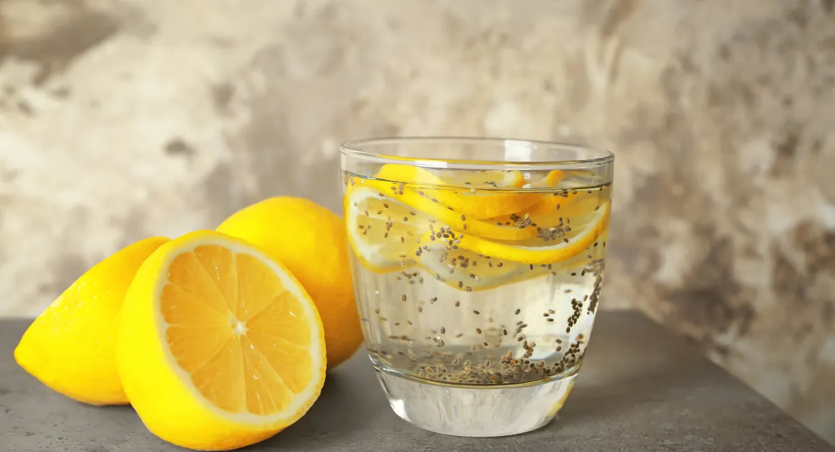 یک لیوان آب، چند عدد لیمو در کنار لیمو و در داخل آب و دانه چیا در داخل لیوان آب