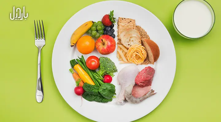 بشقاب حاوی میوه، سبزیجات، کربوهیدرات و پروتئین در کنار یک لیوان شیر روی زمینه سبز